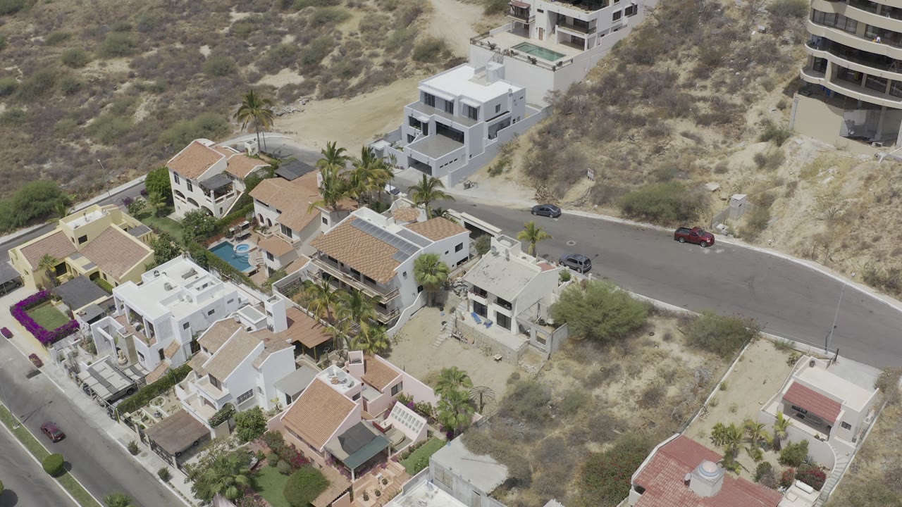 Aerial frame in Los Cabos, Mexico showing modern white homes, nearby tan-roofed houses, small pools, palms, dry hillside brush, a paved road, and no visible people.