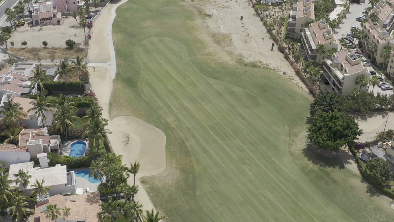 Aerial view of a golf fairway beside homes, pools, palms, and sand bunkers.