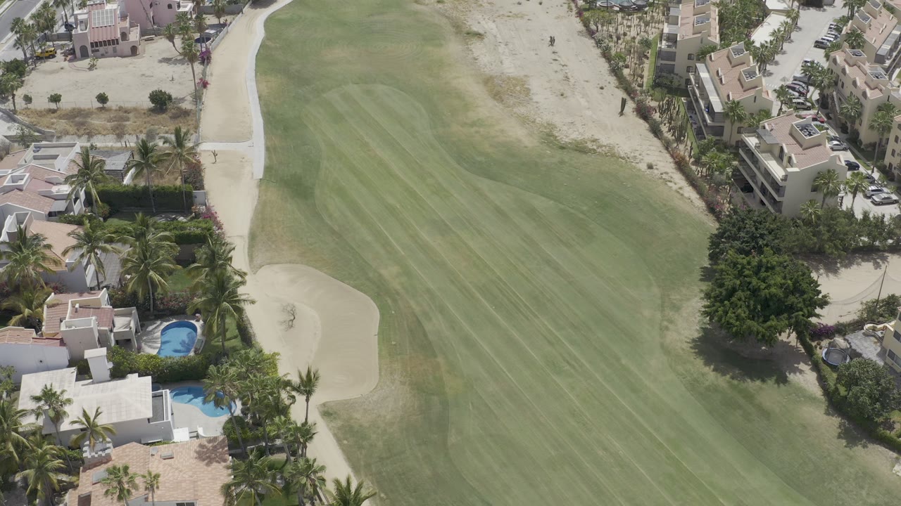 Aerial Los Cabos frame showing the fairway stripes running past a light sand bunker, walled homes, palms, small pools, and dry landscaping with no visible people.