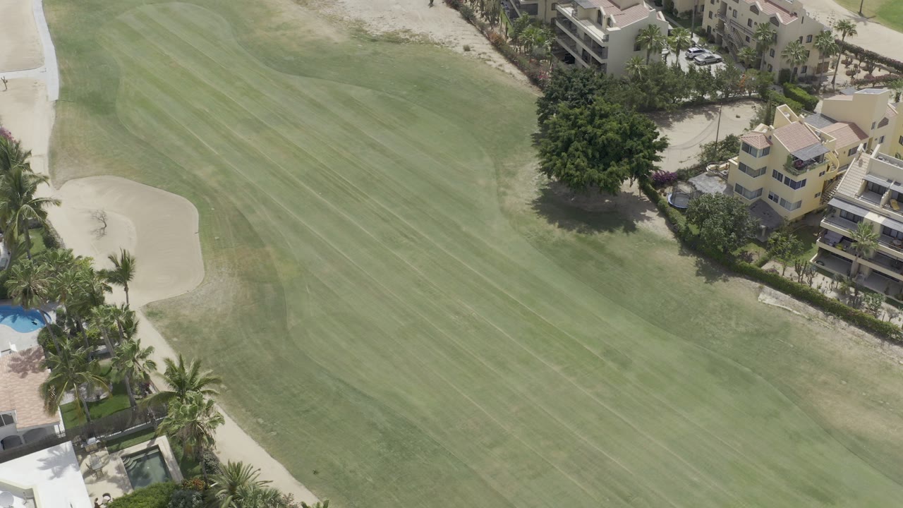 Aerial view of green fairway, sand trap, condos, palms, and pools.