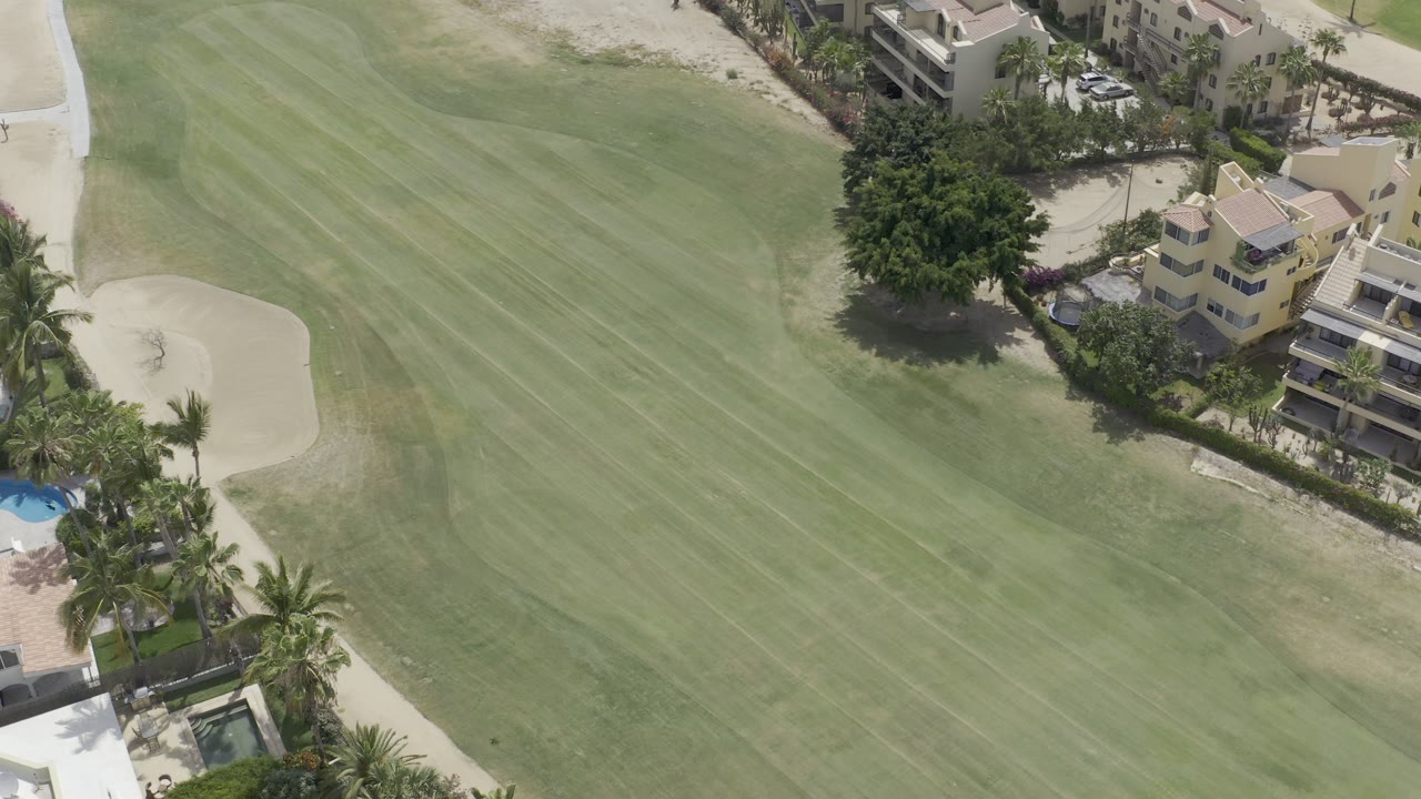 Aerial view in Los Cabos, Mexico, with the green field filling most of the frame and condos, palms, a sand bunker, and pool areas along the upper and lower edges, with no visible people.