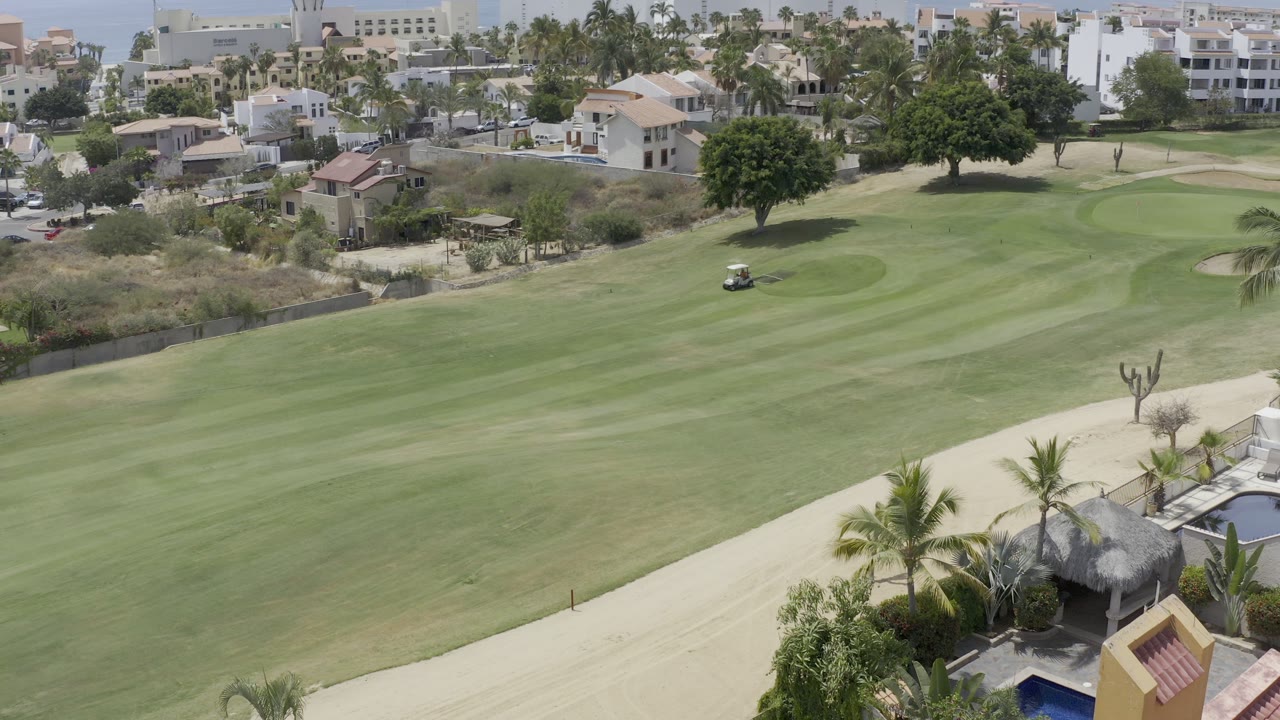 Los Cabos fairway view with a small golf cart in the midground, townhouses beyond the lawn, palms at the lower edge, and no visible people.