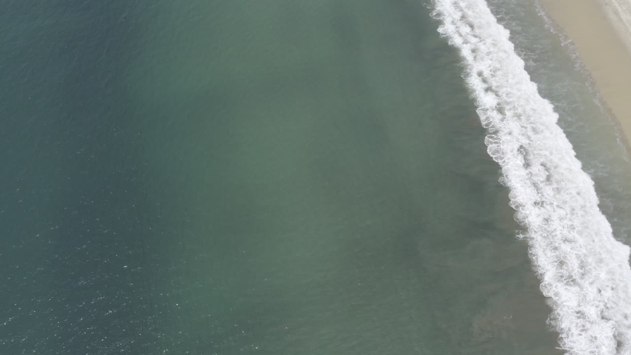 Opening aerial frame over Los Cabos, Mexico water, with blue-green ocean on the left, white surf angled along the right, pale sand at the edge, and no visible people.