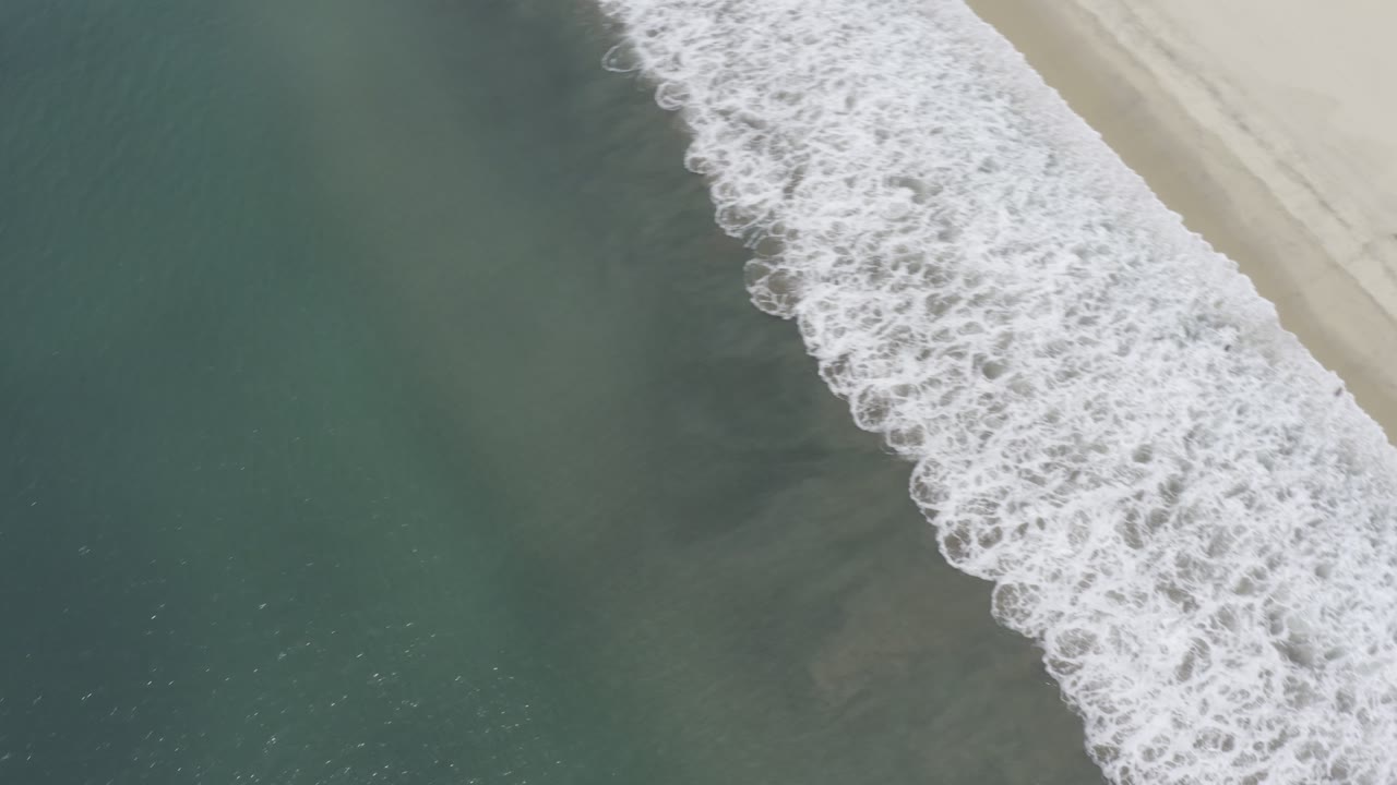 Aerial frame in Los Cabos, Mexico, showing a broad white wave breaking below a narrow strip of pale sand with no visible people.
