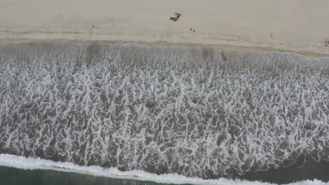 Straight-down aerial view of waves reaching a pale Los Cabos beach, with small people visible near the upper sand at the start of the clip.