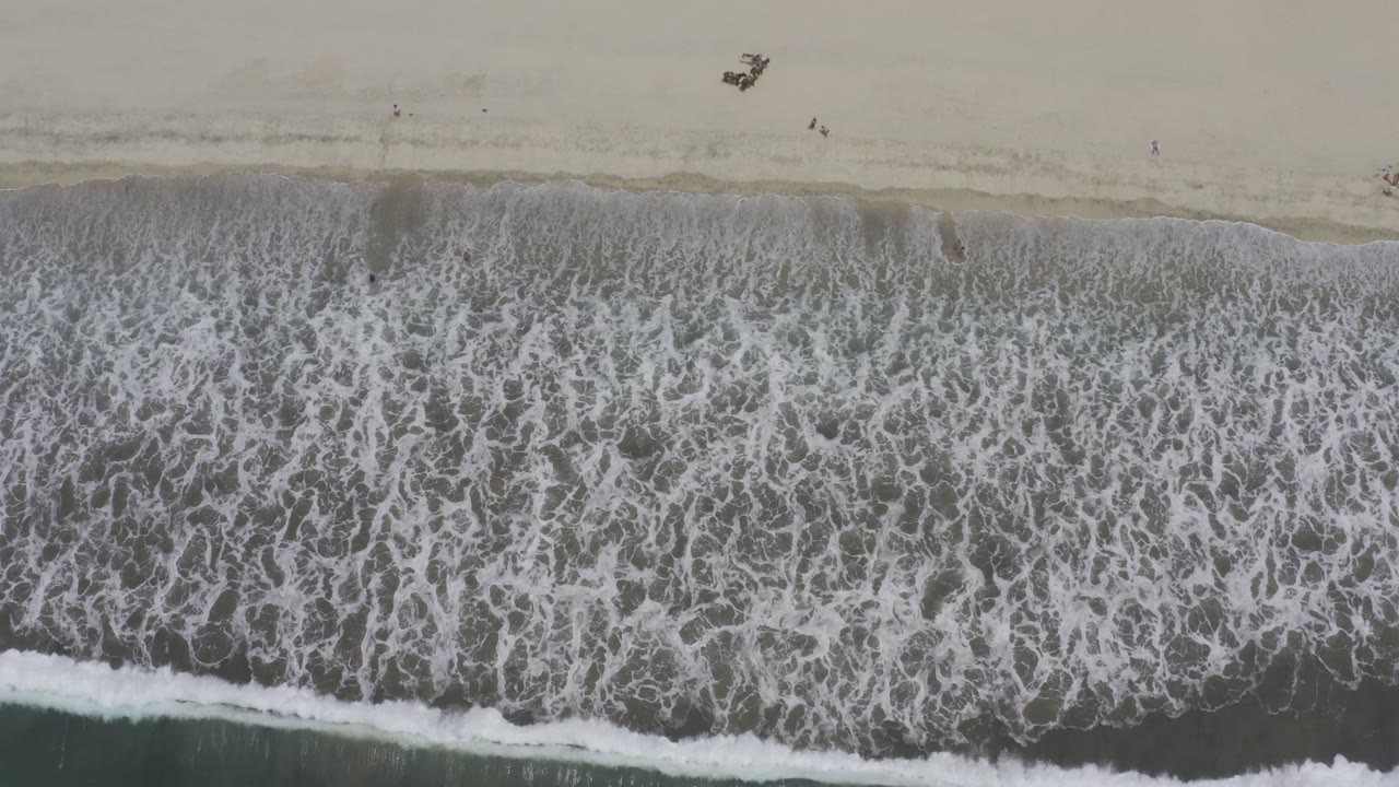Overhead Los Cabos shoreline view with foamy water spread across the lower frame and tiny beachgoers spaced along the upper sand.