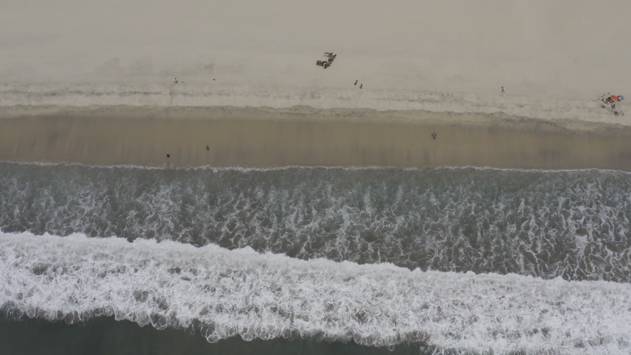 Top-down Los Cabos beach view with gray-green waves and white foam reaching a beige shoreline, with small beach objects near the upper sand and no visible people.