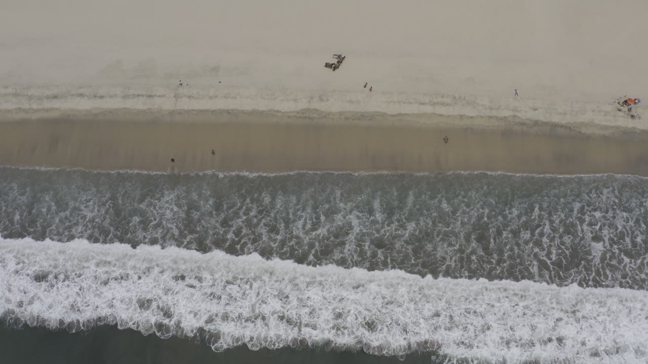 Near-vertical Los Cabos shoreline frame with a wide band of foamy surf below, darker wet sand at center, pale dry sand above, and tiny indistinct objects on the beach.