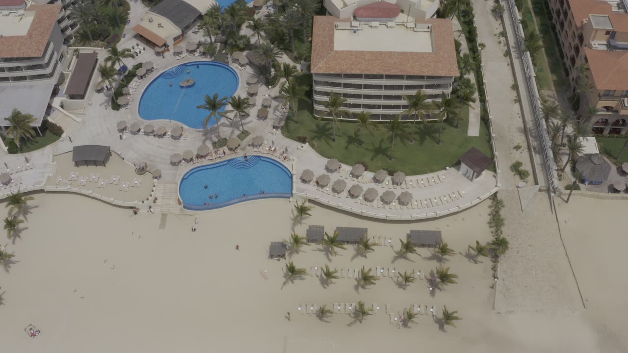 Overhead drone frame in Los Cabos showing two blue resort pools, a sandy beach edge, palm trees, umbrellas, lounge chairs, and hotel buildings, with no visible people.