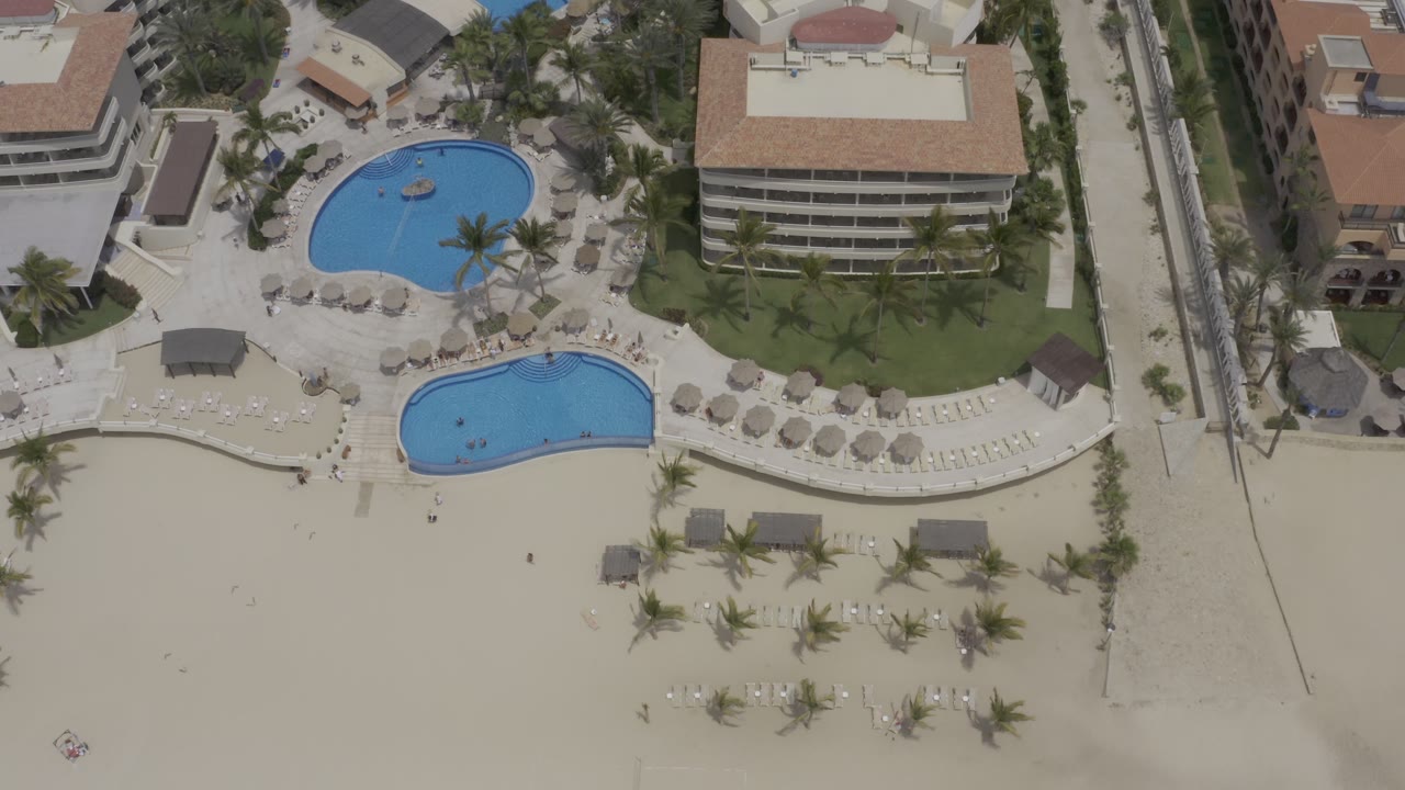 Los Cabos overhead view with two bright pools, tan-roofed buildings, a narrow road along the right side, palms, umbrellas, and beach frontage, with no visible people.