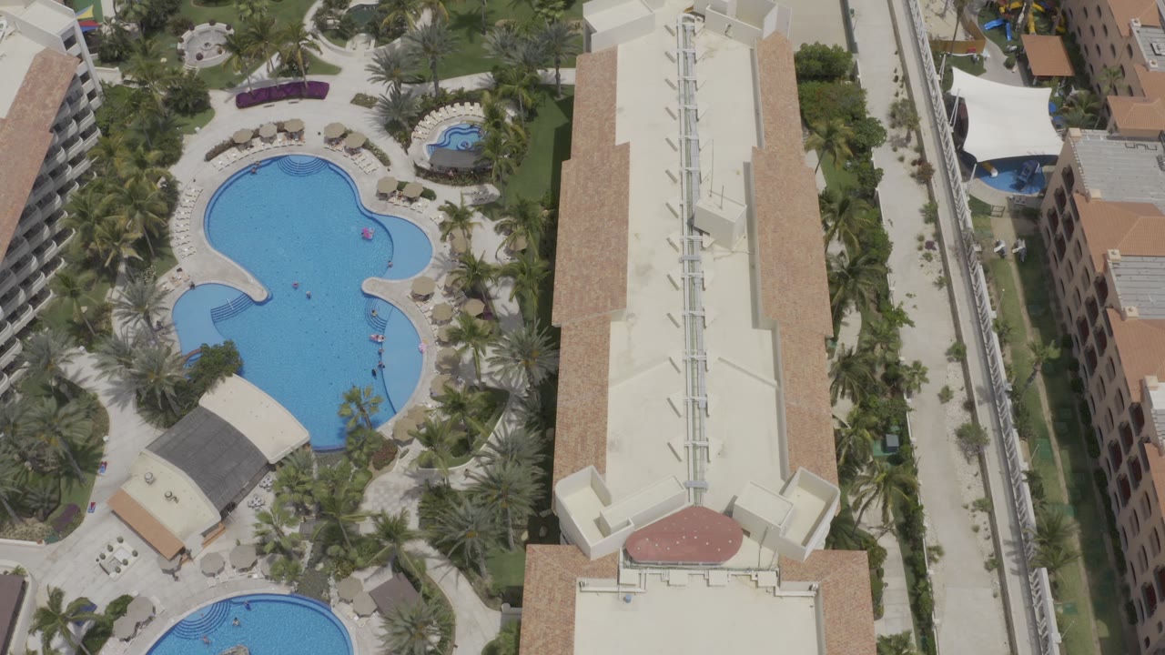 Aerial view in Los Cabos, Mexico, showing two blue curved pools, palm landscaping, a long white roof, nearby vacation buildings, and tiny people visible in the water.