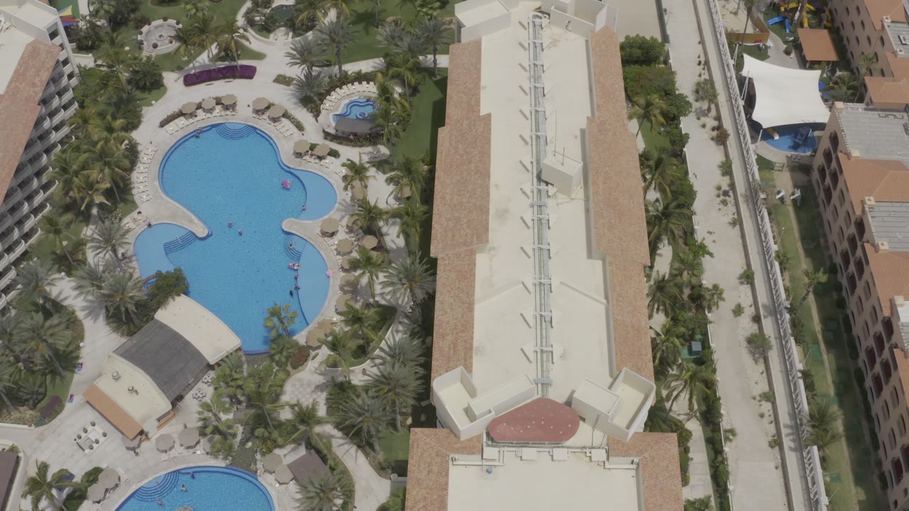 Overhead Los Cabos resort view with a large bright blue pool centered among palms, walkways, lounge areas, tall buildings, and small people visible in the pool.