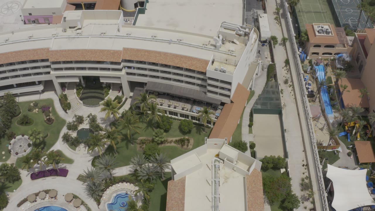 High aerial frame over Los Cabos, Mexico, showing a curved multi-level building, palm courtyard, rooftops, a narrow green-roofed passage, and a sport court with no visible people.