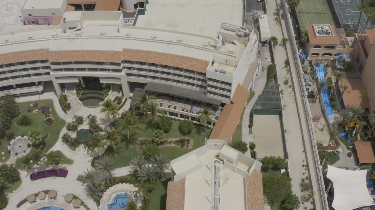 Aerial frame in Los Cabos, Mexico, centered on the curved building and planted courtyard, with adjacent pale rooftops, sport court markings, and no visible people.