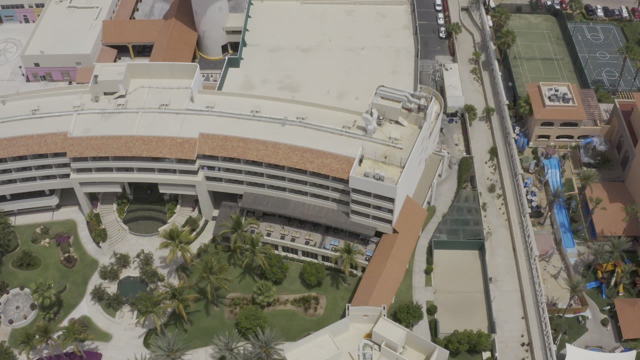 Top-down view of a large pale roof, curved building face, courtyard, and sports area.