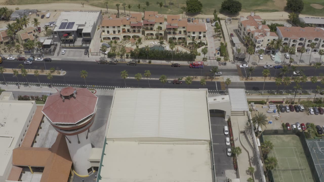 Aerial frame in Los Cabos, Mexico with the road centered, parked cars along lots, light-colored villas, palm trees, and a green fairway behind them, with no visible people.