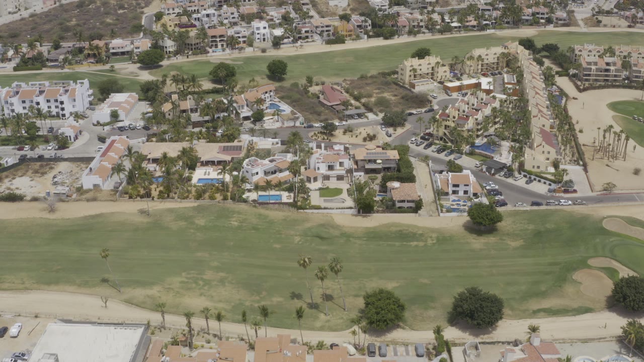 High-angle Los Cabos aerial view with golf fairway, sand traps, villas, roads, palm trees, dry hillside, and no visible people.