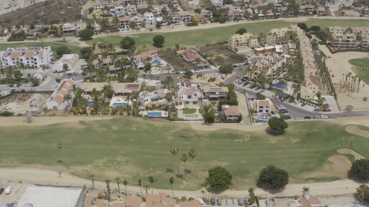 Los Cabos aerial frame showing green fairway in the foreground, clustered homes and apartments beyond it, and dry slopes with no visible people.