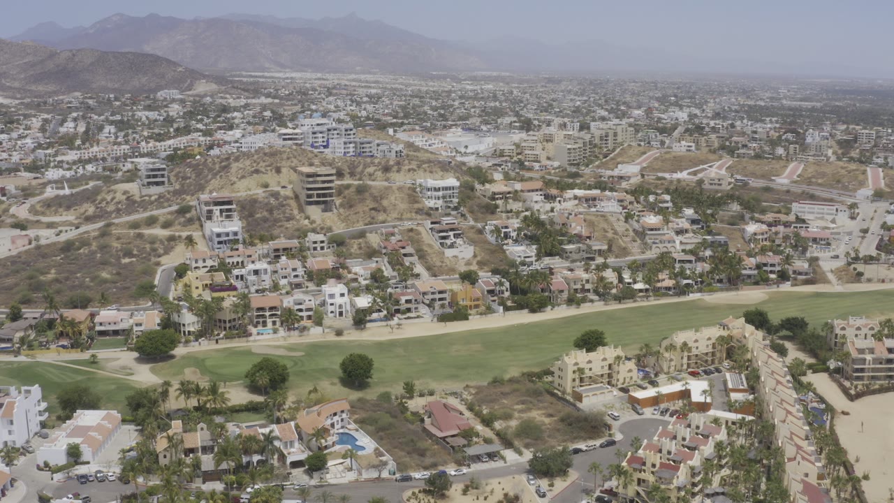 Aerial view over Los Cabos, Mexico, with a green fairway in the foreground, hillside homes and roads in the middle distance, hazy mountains beyond, and no visible people.