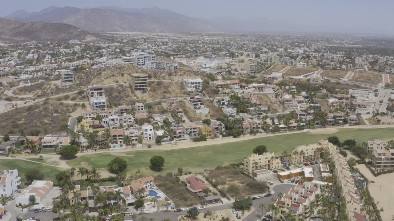 Wide Los Cabos aerial with residential blocks, pale hillside roads, a green golf-course strip across the lower frame, dry terrain, and no visible people.
