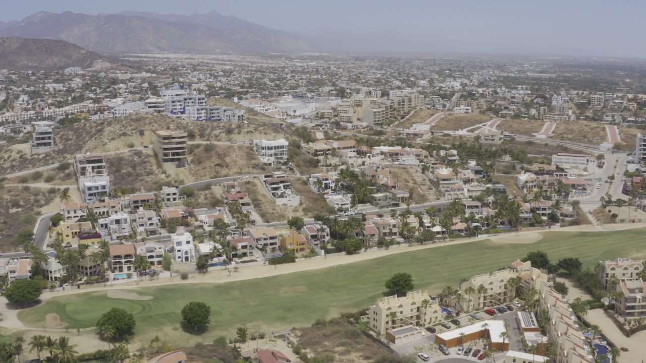 Aerial view of green fairway, hillside homes, roads, city blocks, and hazy mountains.