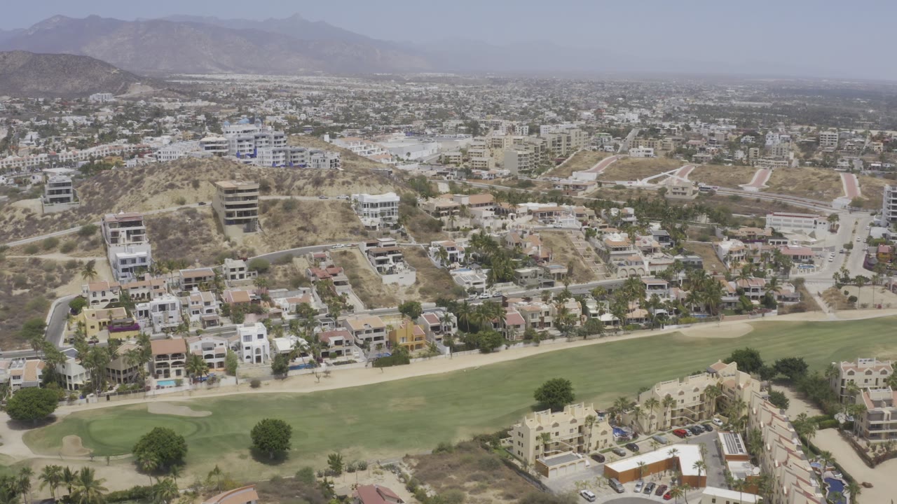 Los Cabos aerial frame with the fairway stretching across the lower image, compact homes and hillside buildings in the middle, distant city blocks, and no visible people.
