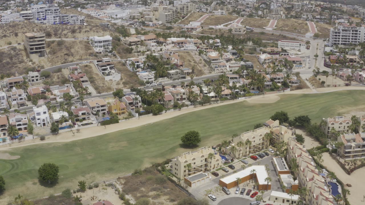 Opening aerial frame over Los Cabos, Mexico, showing a green fairway, sand bunker, residential streets, hillside buildings, and no visible people.
