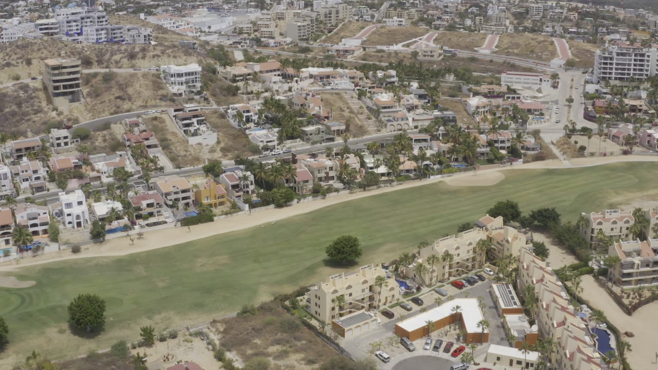 Second aerial frame in Los Cabos, Mexico, with the fairway spanning the lower half, villas and roads across the center, hillside construction above, and no visible people.