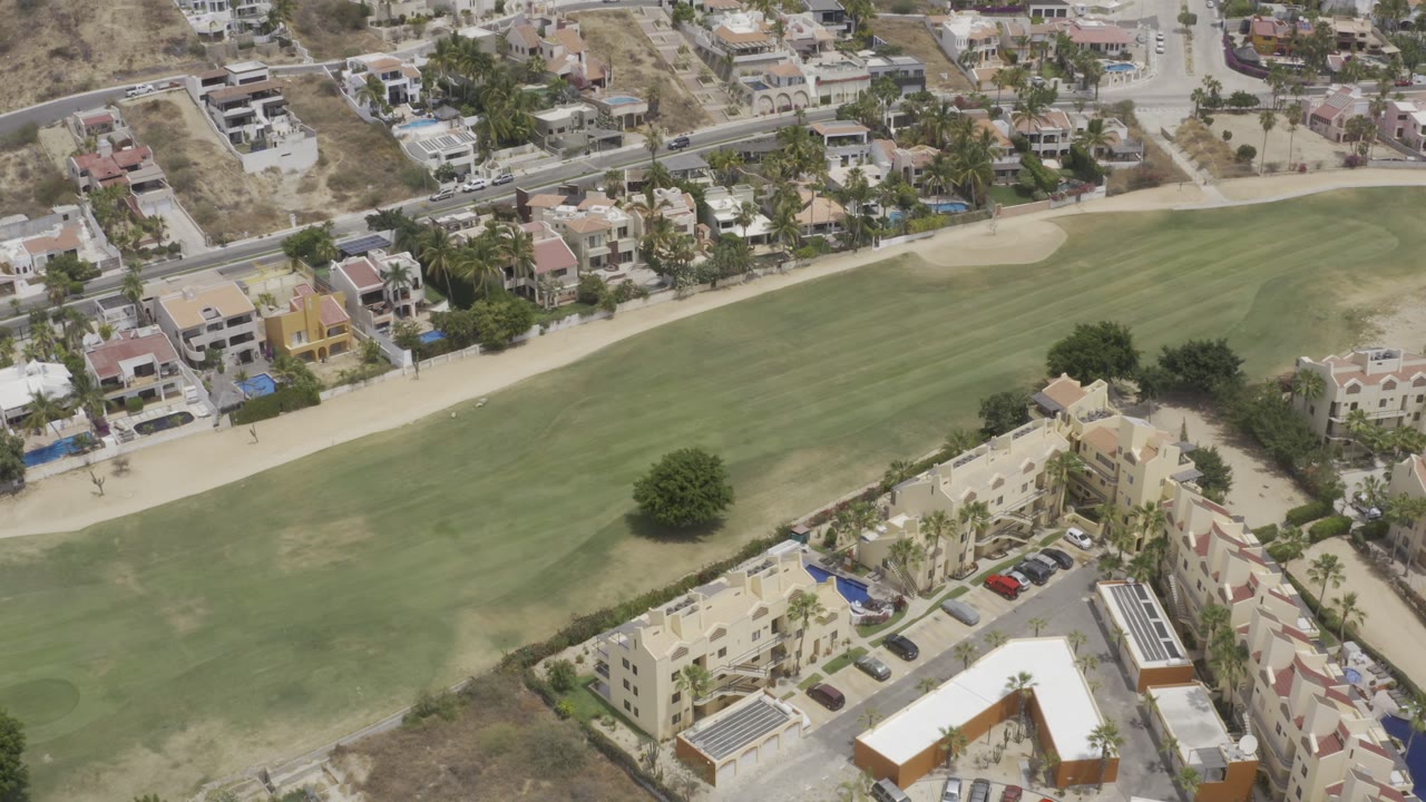 Los Cabos aerial frame with the fairway centered, a small sand trap near the upper edge, homes and parked cars beyond it, and no visible people.