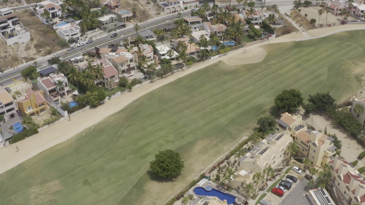 High aerial frame over Los Cabos, Mexico, showing a broad green fairway, pale sand bunker, roadside homes, parked cars, palms, and no visible people.