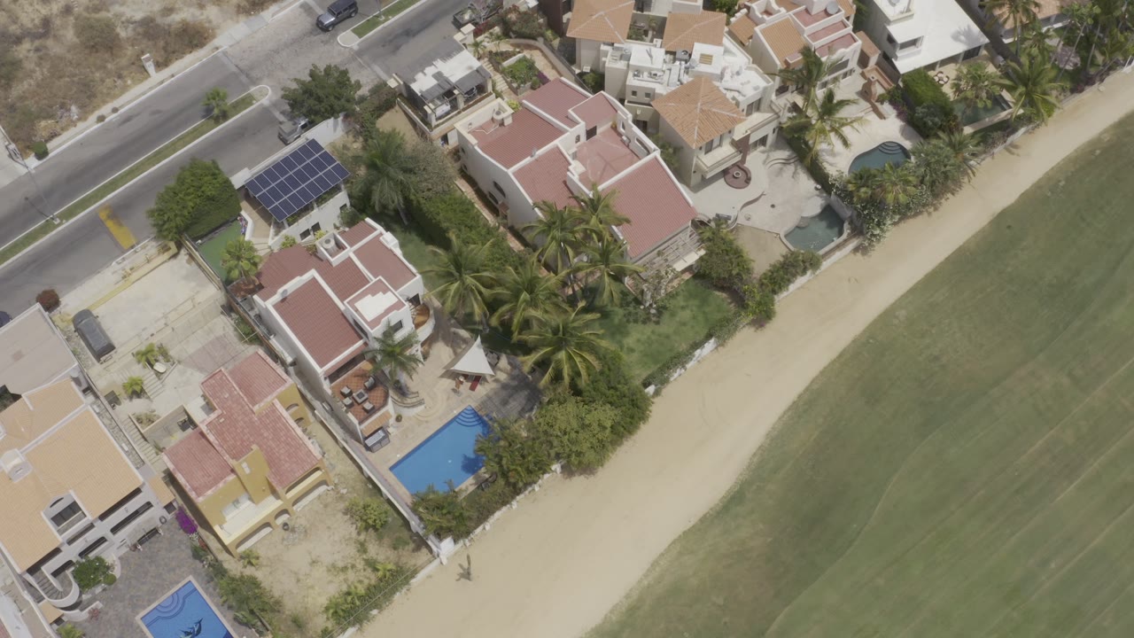 Overhead Los Cabos view with homes shifted farther across frame, blue pools, palm gardens, a roadside curb, green ground, and no visible people.