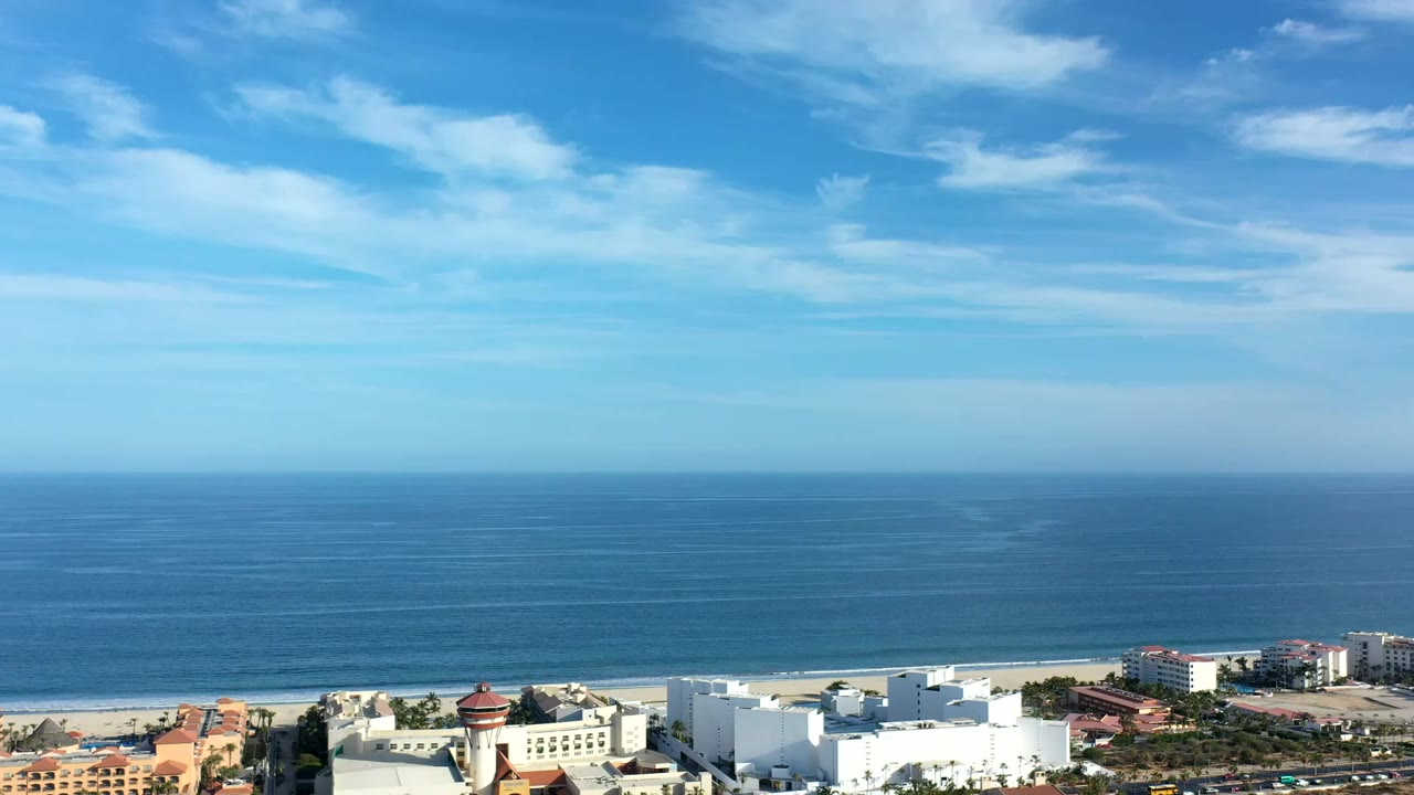 Opening frame of a Los Cabos aerial view with blue ocean, a thin beach line, waterfront buildings at the bottom edge, streaked clouds, and no visible people.