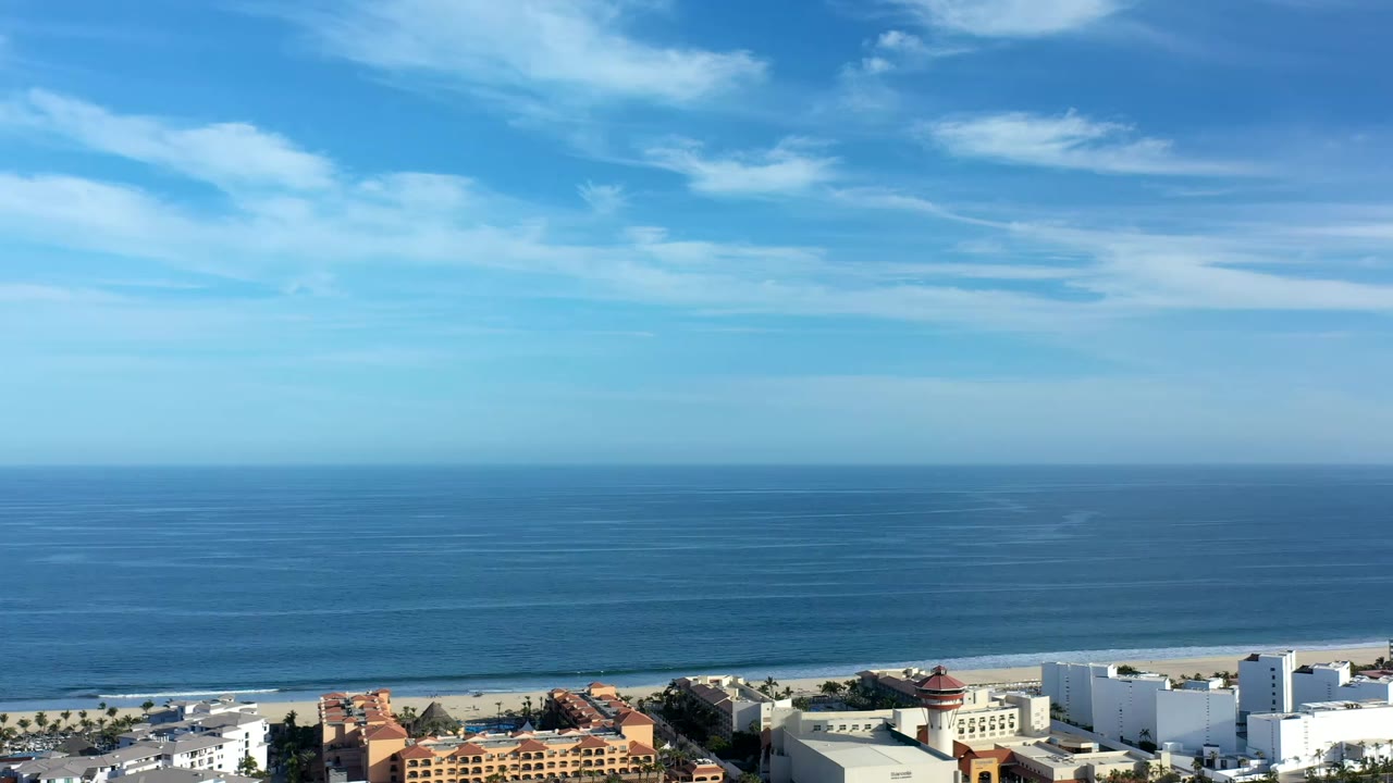 Los Cabos coastline from above with the ocean horizon centered, low surf at the beach, and terracotta and white buildings visible near the lower frame, with no visible people.