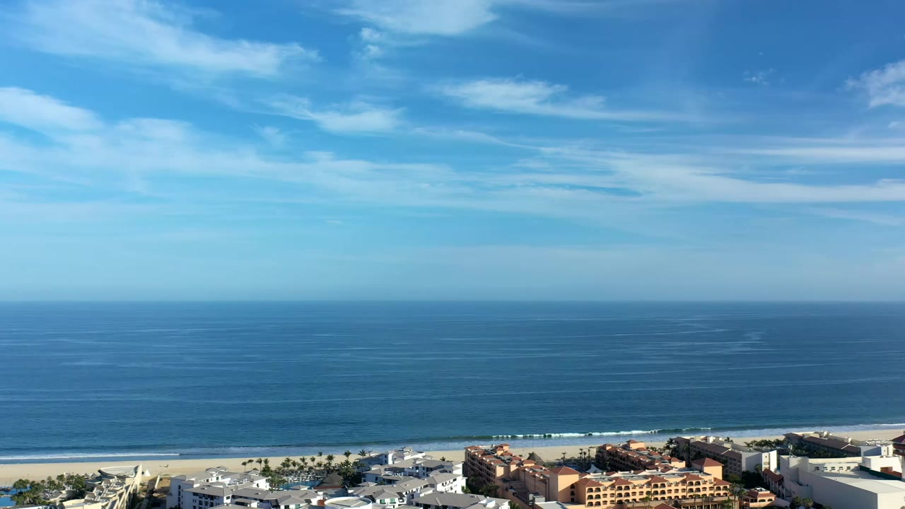 Los Cabos beach frontage sits below a broad blue ocean and level horizon, with low buildings, palms, light cloud cover, and no visible people.