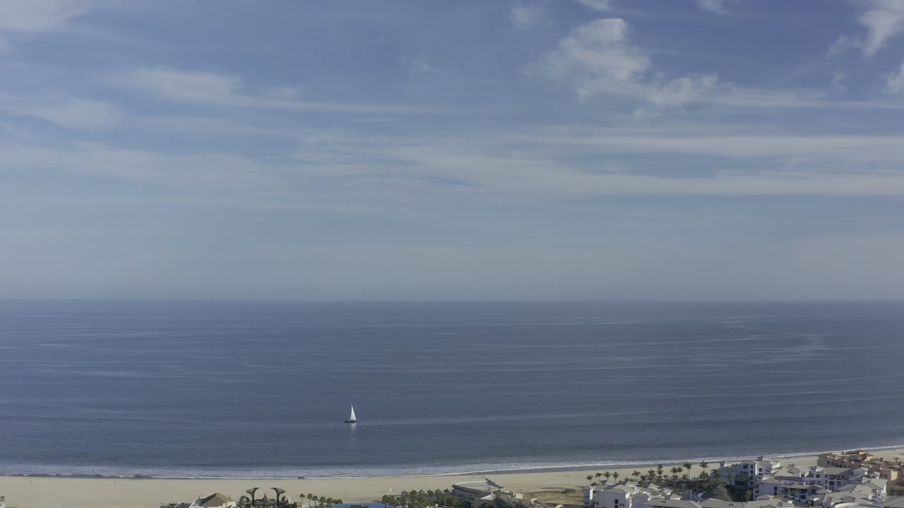 Aerial view in Los Cabos, Mexico, showing a blue ocean horizon, pale beach, low shoreline buildings, a small white sailboat offshore, and no visible people.