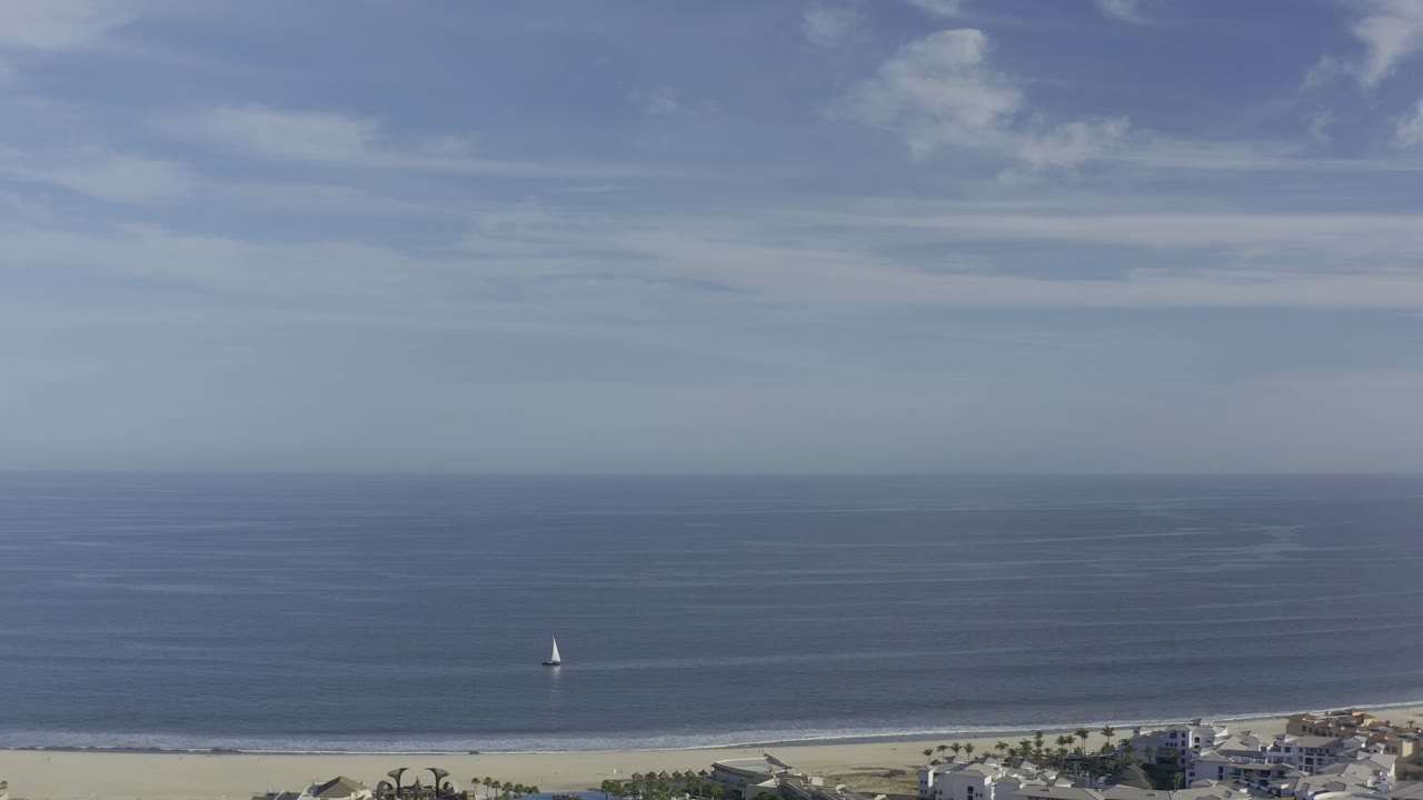 Aerial Los Cabos coastline view with calm blue water, a high horizon, thin surf lines, a small sailboat left of center, beachfront structures, and no visible people.