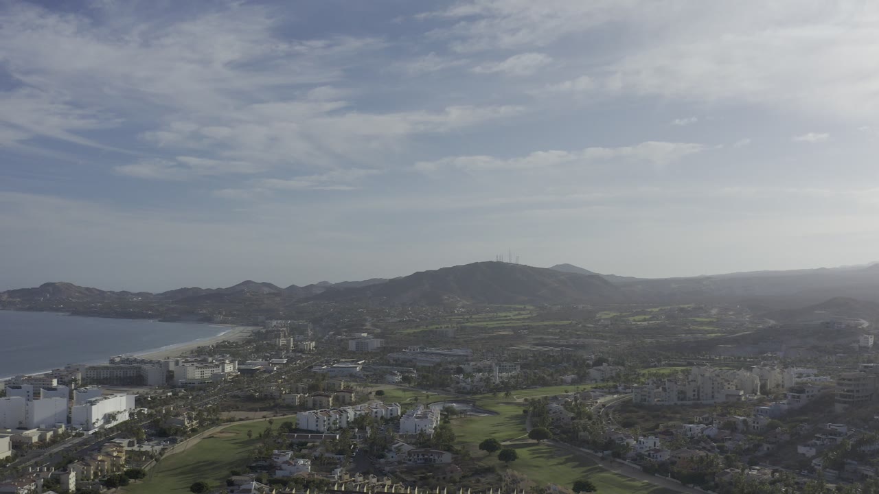 Wide aerial view over Los Cabos, Mexico, showing a bay at left, light resort buildings below, green grounds, dark hills, a hazy horizon, and no visible people.