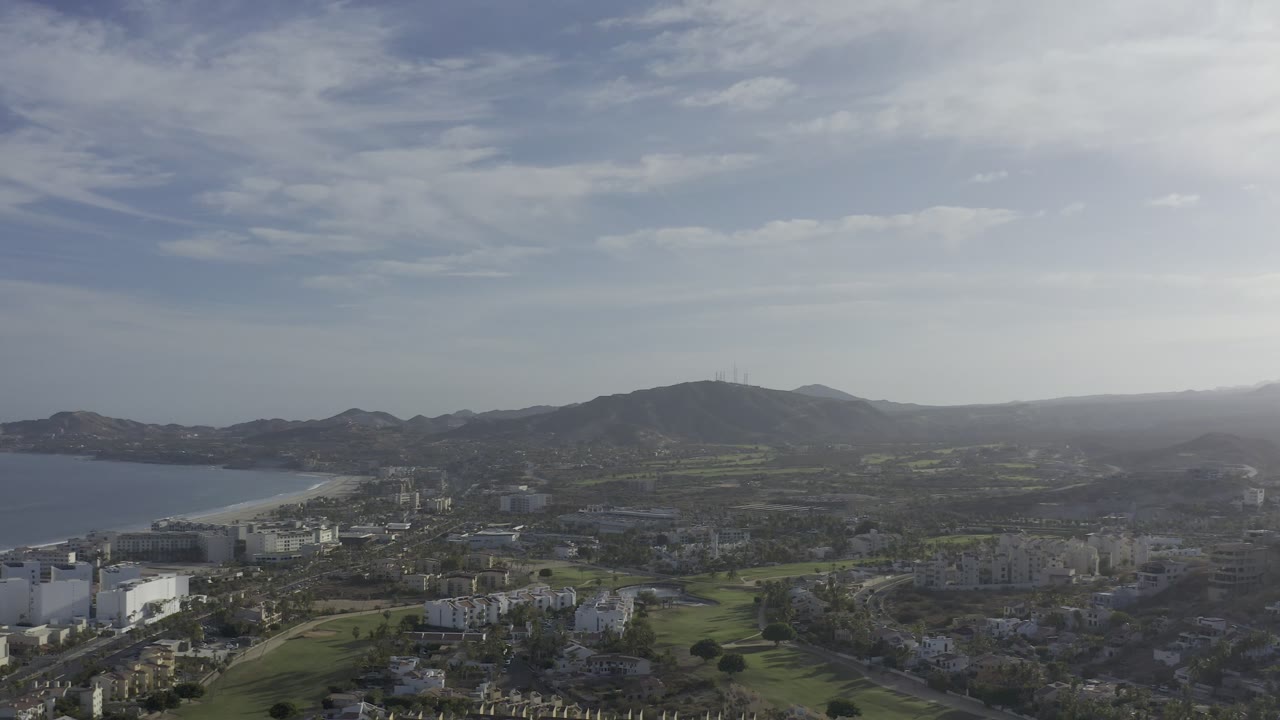 Los Cabos aerial frame with the bay sitting left of center, low buildings and green grounds in the foreground, ridged hills across the middle, and no visible people.