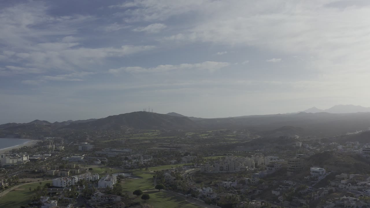 Aerial view over Los Cabos, Mexico, showing a hazy town, green terrain, dark hills, distant ridgelines, and no visible people.