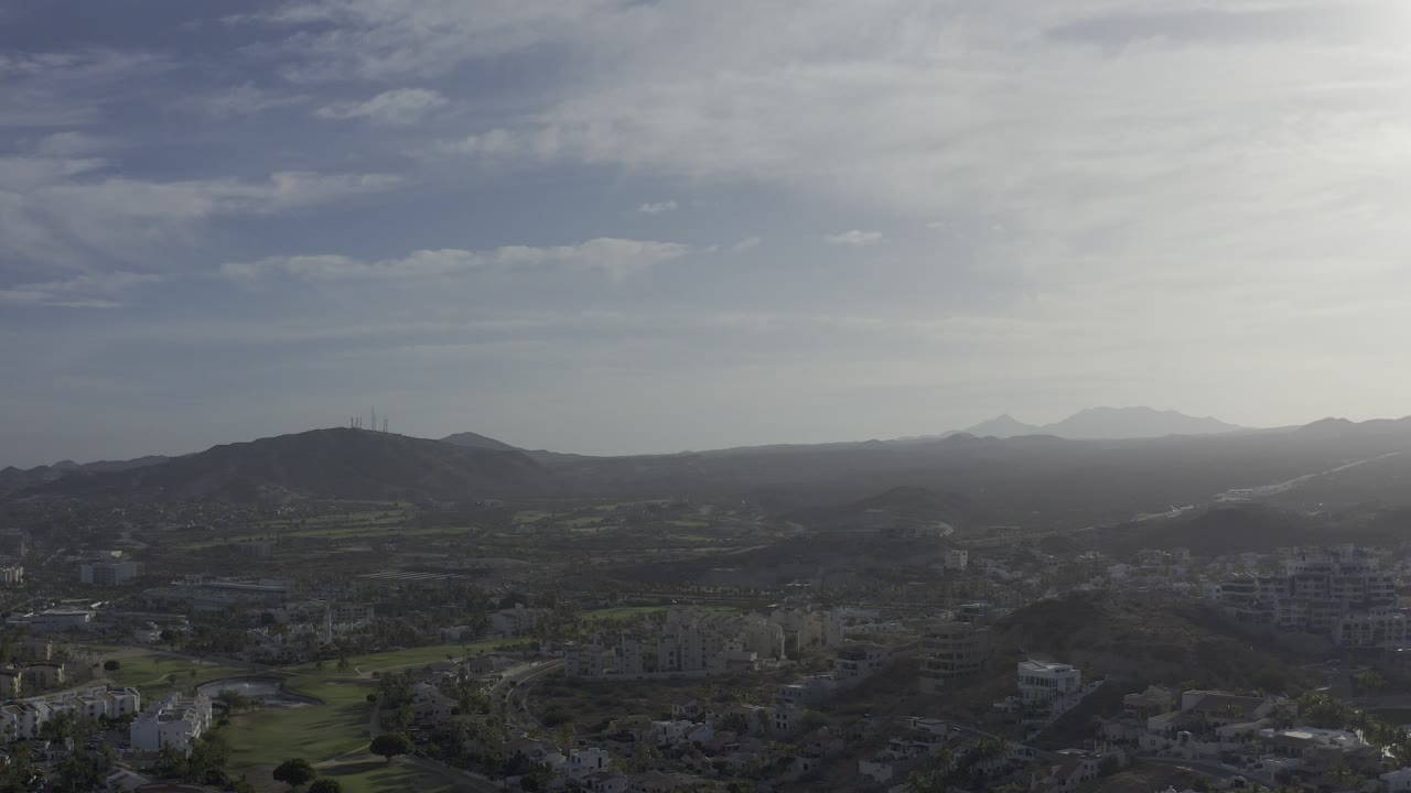 A wide aerial view over Los Cabos shows clustered buildings below, dark hills across the midground, distant mountains, bright haze, and no visible people.