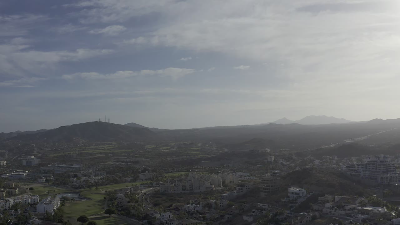Los Cabos appears from above with low-rise buildings in the foreground, green terrain patches, a dark ridge line, distant mountain shapes, and no visible people.