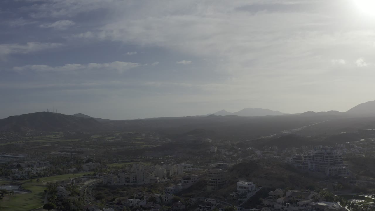 Wide aerial-style view over Los Cabos, Mexico, with a road crossing dark hills, scattered buildings below, distant mountains on the horizon, bright glare at upper right, and no visible people.