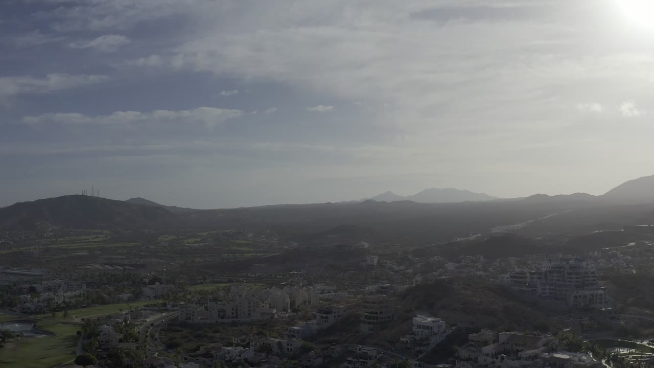 Backlit Los Cabos landscape with the road faintly visible through the midground, dark hillside shapes, pale mountain ridges in the distance, bright sky haze, and no visible people.