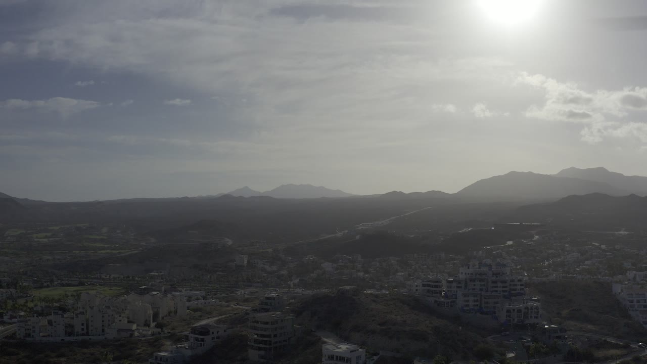 Aerial view over Los Cabos, Mexico with dark foreground buildings, rolling hills, distant mountain silhouettes, bright sun glare, and no visible people.