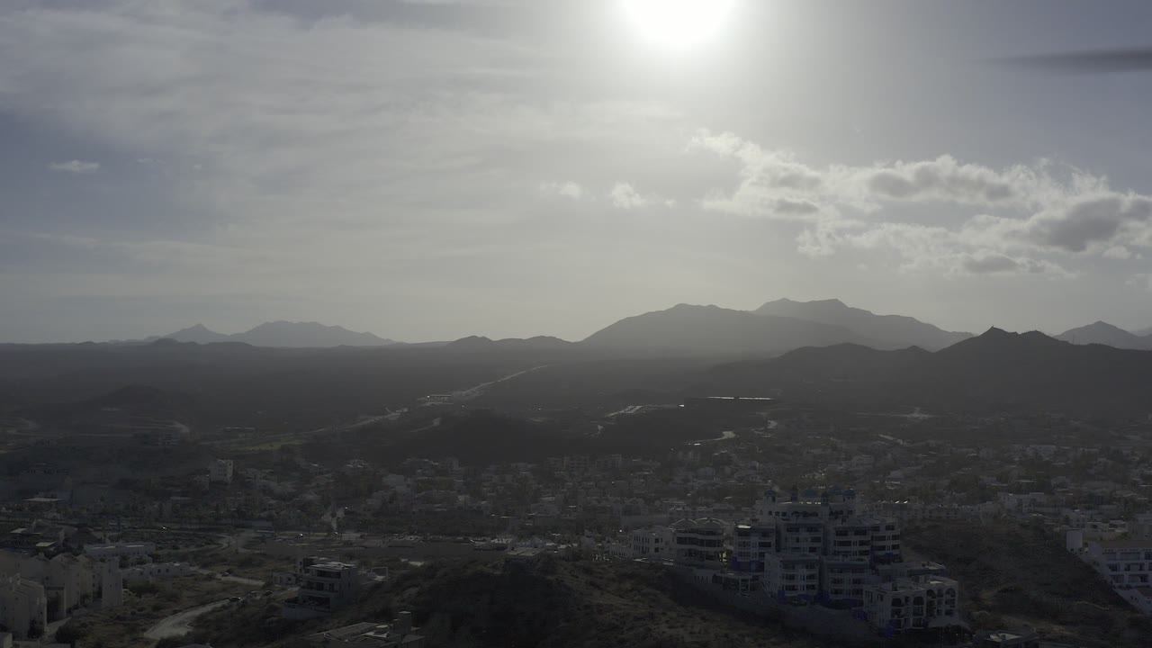 Opening frame of Los Cabos, Mexico from high above low buildings and hills, with a hazy mountain horizon under a bright sun and no visible people.