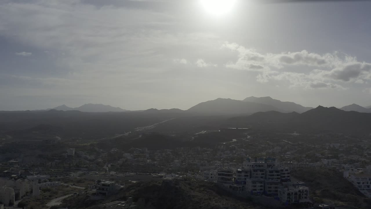 Los Cabos aerial frame with roads and clustered buildings below, dark scrub hills in the middle distance, mountains across the horizon, and no visible people.