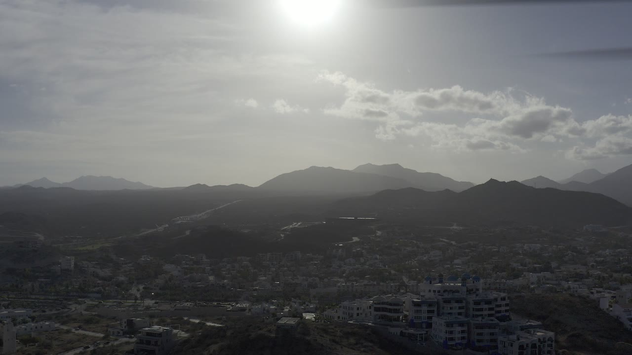 Los Cabos aerial frame with a hazy urban area below layered mountains, bright overhead sun, thin clouds, and no visible people.