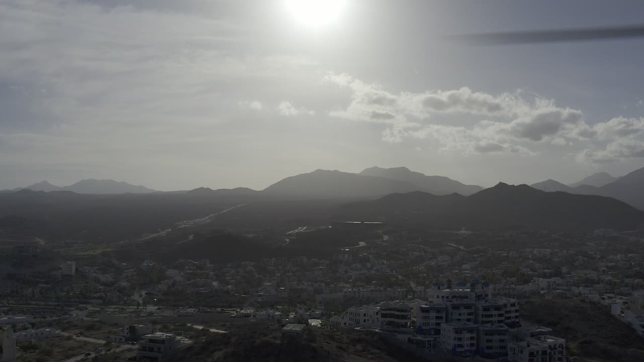 Los Cabos cityscape appears under strong backlight, with road lines crossing the valley, dark mountain silhouettes, and no visible people.