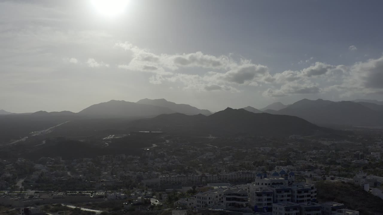 Los Cabos mountain horizon under strong sun glare, with low buildings and dark hills in the lower frame and no visible people.