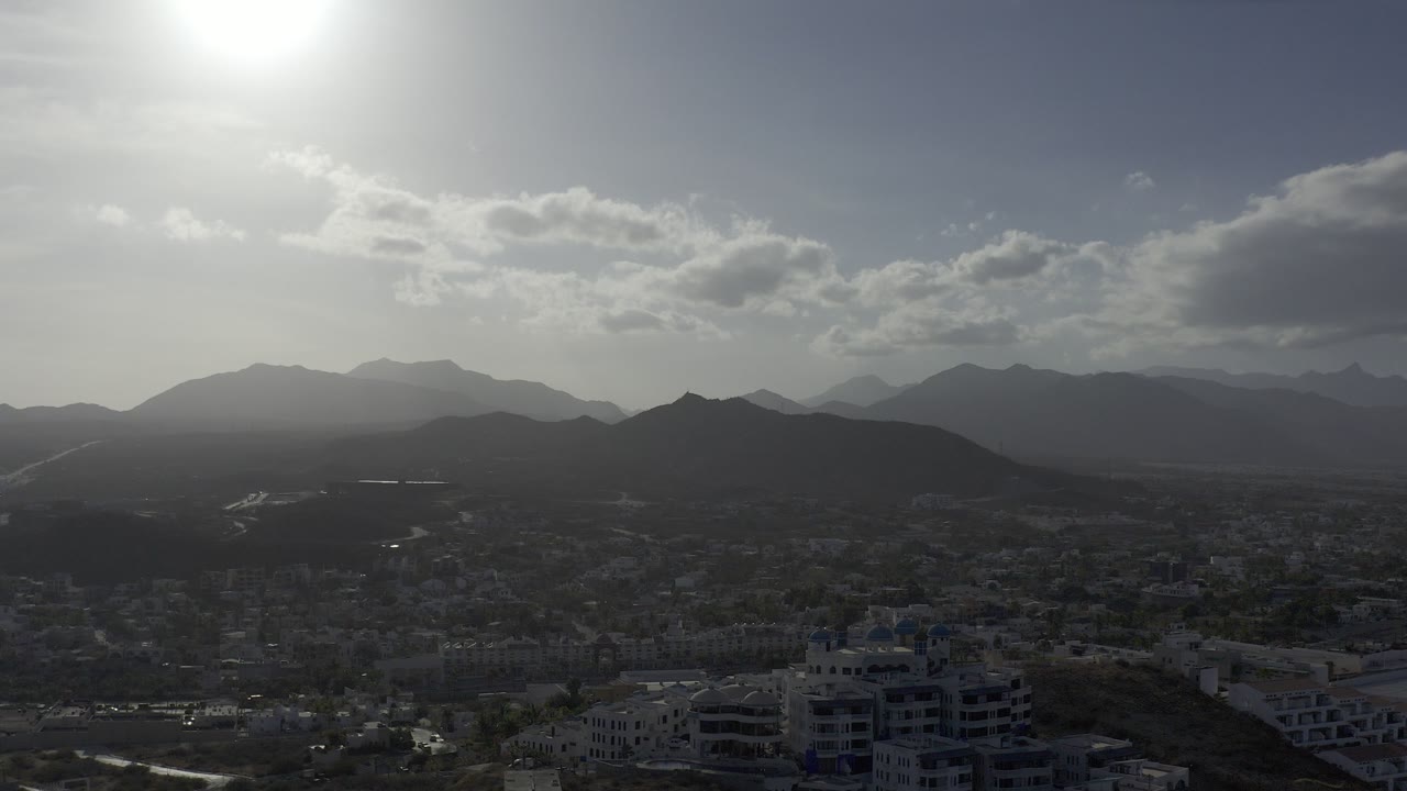 Wide Los Cabos, Mexico skyline at sunrise with the sun flaring near the upper left, layered mountains across the horizon, dark buildings below, and no visible people.